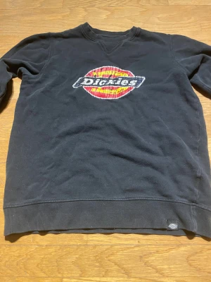 Svart sweatshirt från Dickies M - Svart sweatshirt från Dickies i storlek medium med stort tryckt logomärke i rött, gult och vitt på bröstet. Tröjan har rund hals, långa ärmar och ribbade muddar vid ärmslut och nederkant. Materialet är mjuk bomull och polyesterblandning.