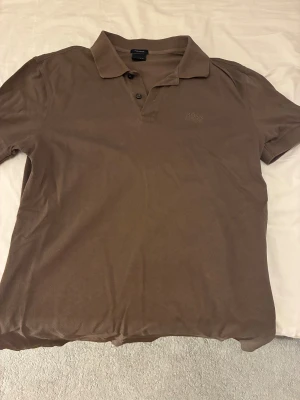 Brun pikétröja från BOSS - Säljer en stilren brun pikétröja från BOSS med klassisk krage och två knappar framtill. Tröjan har diskret BOSS-brodyr på bröstet och är tillverkad i mjuk bomull. Perfekt för en clean och avslappnad look.