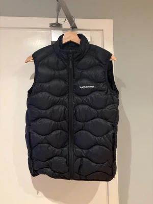 Svart dunväst från Peak Performance, strl 170 - Säljer en svart dunväst från Peak Performance med hög krage och dragkedja framtill. Västen har quiltad design och två fickor med dragkedja. Perfekt för lager på lager och riktigt snygg till hoodie eller tröja. Strl 170