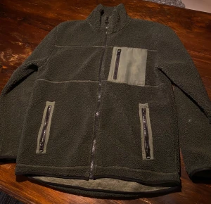 Mörkgrön teddyjacka från 157 - Mysig mörkgrön teddyjacka från 157 med hög krage och tre praktiska fickor med dragkedja, varav en på bröstet. Jackan har en loose passform och är tillverkad i fleece med detaljer i syntetmaterial. Perfekt för kyliga dagar och chill stunder.