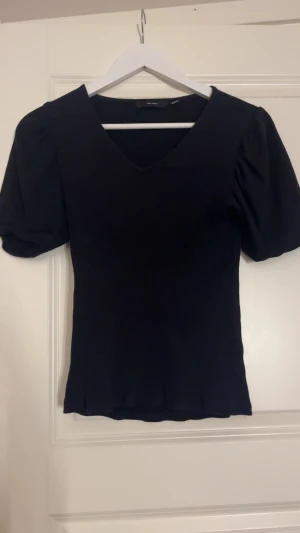 Svart v-ringad topp från Vero Moda - Svart topp från Vero Moda med v-ringning och puffiga korta ärmar. Modellen är figurnära med lätt ribbad struktur och mjukt material som sitter snyggt. Perfekt för dig som gillar stilrena och enkla plagg med en twist.