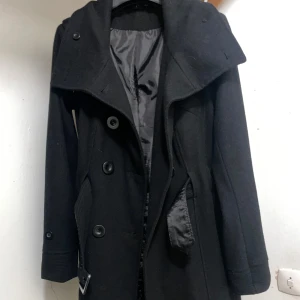 Svart kappa med bälte och stora slag - Snygg svart kappa med breda slag och dubbelknäppning. Jackan har ett matchande bälte i midjan och stora knappar framtill. Fodrad insida och långa ärmar ger en clean och stilren look. Perfekt för dig som gillar klassisk stil med edge. Går ner till knäna, jag är 168