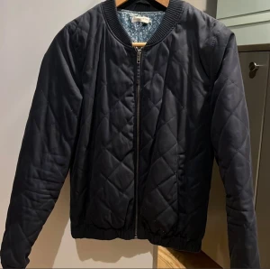 Marinblå quiltad bomberjacka JDY S - Marinblå quiltad bomberjacka från Jacqueline de Yong i storlek S. Jackan har ribbade muddar, dragkedja framtill och ett stilrent, rutigt quiltmönster. Två fickor fram. Blommigt foder uppe vid nacken. Längd ca 55cm.