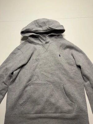 Grå hoodie från Polo Ralph Lauren - Snygg grå hoodie från Polo Ralph Lauren med klassisk huva och magficka. Tröjan har ett litet mörkblått Polo-logo broderat på bröstet och är tillverkad i mjuk bomullsmix. Den passar S.