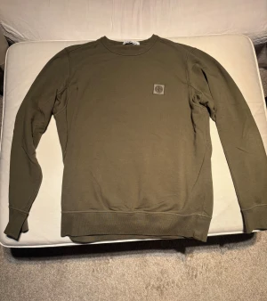 Stone Island sweatshirt Olivgrön - Säljer en olivgrön sweatshirt från Stone Island med klassisk rund hals och ribbade muddar. Tröjan har lång ärm och en liten patch med loggan på bröstet. Perfekt för dig som gillar stilrena och enkla plagg med streetkänsla.