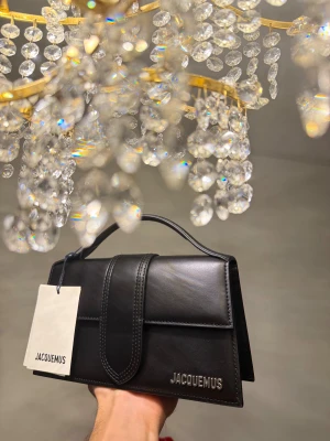 Jacquemus the large bambino  - Snygg svart handväska från Jacquemus med stilren, rektangulär form och silverfärgad logga. Väskan har ett topphandtag och medföljande axelrem med metallspännen. Perfekt för dig som gillar design och vill ha något som sticker ut.