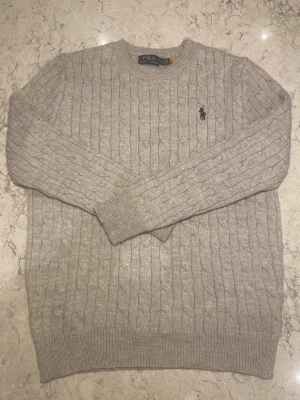 Grå stickad tröja Polo Ralph Lauren - Snygg grå stickad tröja från Polo Ralph Lauren i klassisk kabelstickad design. Tröjan har rund hals, långa ärmar och den ikoniska broderade loggan på bröstet. Perfekt för dig som gillar stilrena och tidlösa plagg med premiumkänsla.