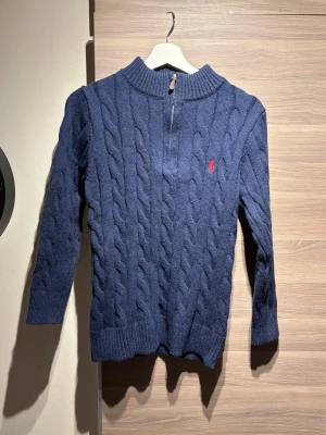Marinblå stickad tröja Polo Ralph Lauren - Säljer en marinblå stickad tröja från Polo Ralph Lauren med klassiskt kabelstickat mönster och halv dragkedja vid halsen. Tröjan har långärmad design, ribbad krage och den ikoniska röda Polo-loggan broderad på bröstet. Storleken är M men passar bra på S också 