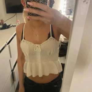 Vit stickad linnetopp med volang - Supersöt vit stickad linnetopp med smala axelband och volang nertill. Toppen har ribbad struktur, liten knytning framtill och croppad passform. Perfekt för sommardagar och enkel att styla med jeans eller kjol.