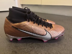 Nike Mercurial Zoom fotbollsskor - Säljer ett par Nike Mercurial Zoom fotbollsskor i metallic koppar med svarta och vita detaljer. Skorna har snörning, hög strumpliknande krage och rosa insida. Yttersulan har rosa och vita dobbar för gräsplan. Snyggt mönstrad ovandel i syntetmaterial.