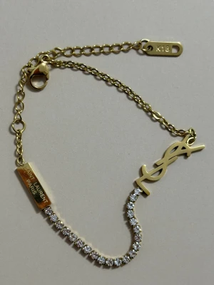 18 k Guldpläterad armband  - Exklusivt armband från Saint Laurent Paris i 18K Guldpläterad med YSL-logga och gnistrande vita stenar. Kedjan är tunn och elegant med justerbar längd och karbinlås. Perfekt för dig som vill ha något lyxigt och stilrent.