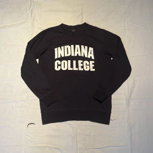Svart college-tröja Indiana College - College-tröja från Smog i svart med vit tryckt text 'INDIANA COLLEGE' på bröstet. Regular fit med rund hals och långa ärmar. Mjuk bomullskänsla och klassisk stil, perfekt för chill dagar eller när du vill ha en enkel look.