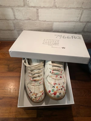 Masion margiela gats  - Maison Margiela  sneakers i vitt skinn med coola färgstänk i rött, blått, orange och svart på tån och sidorna. Klassisk låg modell med snörning och gummisula. Tillverkade i lamm- och kalvskinn, med en unik konstnärlig vibe som sticker ut.