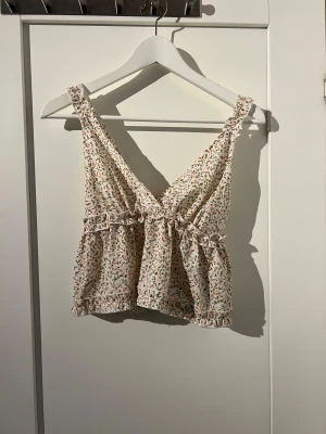 Blommig volangtop med v-ringning - Supersöt blommig linnetopp i vitt med små rosa och gröna blommor, volangdetaljer och v-ringning. Toppen har breda axelband och är lätt utsvängd nertill. Perfekt för varma dagar och ger en romantisk vibe.