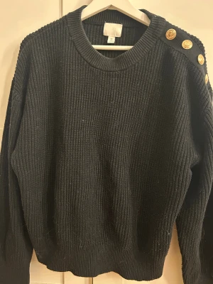 Svart stickad tröja med guldknappar H&M - Svart stickad tröja från H&M i storlek L. Tröjan har grov ribbad struktur och rund hals. På ena axeln finns fyra snygga guldknappar som ger en cool detalj. Perfekt för dig som gillar enkel men stilren design.