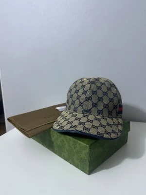 Gucci beige keps med GG-mönster - Snygg beige keps från Gucci med klassiskt blått GG-mönster över hela kepsen och en blå-röd rand på sidan. Justerbar rem bak i blått läder. Kepsen är tillverkad i bomull och har en stilren, rundad form som ger en lyxig vibe.