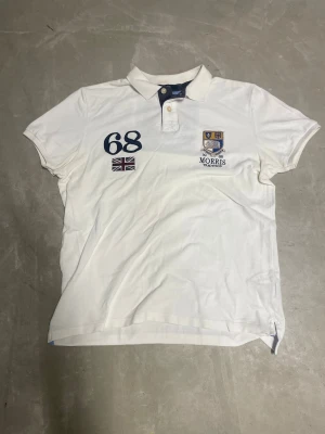 Vit pikétröja Morris Tradition - Snygg vit pikétröja från Morris Tradition med broderad logga, siffran 68 och brittisk flagga på bröstet. Klassisk krage med två knappar och blå detaljer på insidan av kragen. Perfekt för dig som gillar preppy stil och vill sticka ut med coola broderier. Storlek L men funkar som M om du föredrar mer loose fit.