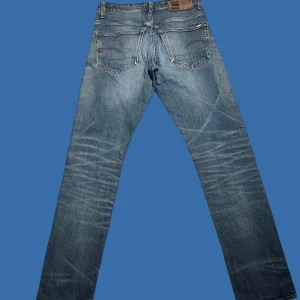 Blå jeans från G-Star RAW - Snygga blå jeans från G-Star RAW med klassisk femficksdesign och läderpatch bak i midjan. Jeansen har en rak passform och coola slitningar framtill som ger en avslappnad vibe. Perfekta för dig som gillar streetstyle och vill ha ett par jeans med karaktär.