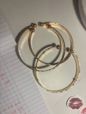 Tre guldiga armband med mönster - Säljer tre snygga guldiga armband i metall med graverade mönster och kulor i ändarna. Armbanden är öppna och har olika detaljerade gravyrer som stjärnor och trianglar. Perfekta för att ge din outfit lite extra bling.