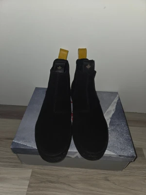 Svarta chelsea boots från Canada Snow - Snygga svarta chelsea boots från Canada Snow i mocka med elastiska paneler på sidorna och en gul hälflik i skinn. Klassisk rund tå och platt sula gör dem både stilrena och lätta att ta på. Perfekta för dig som vill ha en clean look med en twist. Helt nya använda 1 gång storlek 42 men passar 43 också.