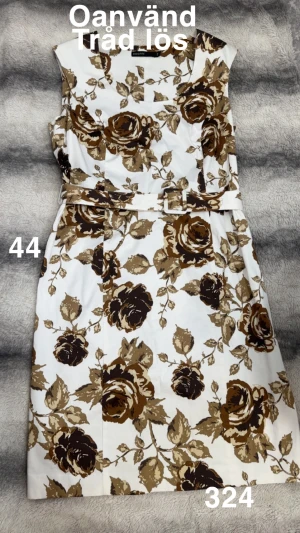 Vit blommig klänning med bälte - Säljer en vit klänning från KappAhl med brunt och beige blommönster. Klänningen är ärmlös, har rund hals och medföljande bälte i midjan. Modellen är knälång och har en klassisk, feminin siluett som passar dig som gillar retrovibbar och tydliga detaljer.🌸Oanvänd. Dock har snöret som håller i skärpet släppt på ena sidan. Storlek 44