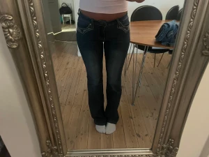 Mörkblå bootcut jeans med broderi - Säljer ett par mörkblå bootcut jeans med snygga broderade detaljer på bakfickorna och kontrastsömmar. Jeansen har låg midja och klassisk femficksdesign. Perfekta för dig som gillar en cool och trendig look.