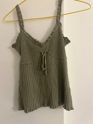 Olivgrön ribbad topp med knyt - Supersöt olivgrön linnetopp med ribbad struktur och volangkant. Toppen har smala axelband, dekorativ knytning framtill och en beige spetsdetalj vid v-rigningen.