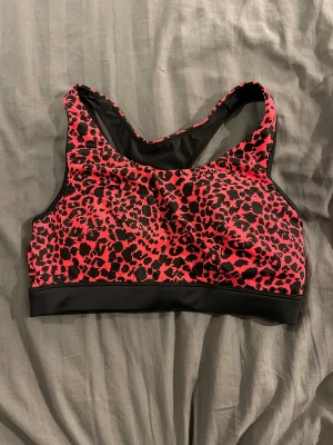 Leopardmönstrad sporttopp i rosa och svart - Sporttopp med coolt leopardmönster i rosa och svart. Modellen är ärmlös med breda axelband och brottarrygg. Svart resår nedtill för extra stöd. Perfekt för dig som vill sticka ut på gymmet eller under träningspasset.