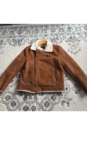 Brun teddyjacka från Burton Menswear - Säljer en brun teddyjacka från Burton Menswear London i storlek medium. Jackan har mjukt vitt teddyfoder, två fickor med dragkedja och en snygg krage. Perfekt för kalla dagar och har en cool, klassisk look med detaljer i syntetmaterial.