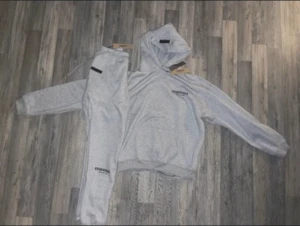 Grå Essentials hoodie med huva - Säljer en ljusgrå hoodie från Essentials med huva och svart logga på bröstet. Hoodien har en klassisk passform, ribbade muddar och är gjord i mjukt bomullsmaterial. Perfekt för en chill och avslappnad stil.