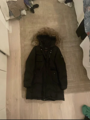 Svart parkas från Hollies med päls - Svart lång parkas från Hollies med snygg fuskpäls på huvan och flera praktiska fickor framtill. Jackan har dragkedja och knappar, samt ett H-emblem på ärmen. Perfekt för kalla dagar med sin varma och stilrena look.