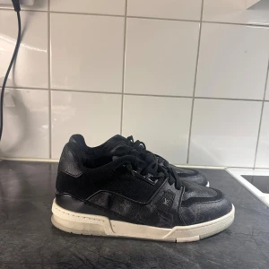 Louis vuitton - Svarta Louis Vuitton sneakers i skinn med klassiskt monogrammönster och vita sulor. Skorna har snörning och rund tå, samt diskreta LV-detaljer på sidan och hälen. Snygg kontrast mellan svart ovandel och vit sula, perfekt för dig som gillar lyxiga streetwear-vibbar.