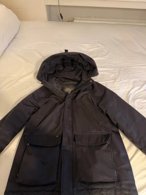 Svart parkajacka med huva från Nobis - Svart parkajacka från Nobis med stor huva och flera praktiska fickor framtill. Jackan har dragkedja och tryckknappar, lång ärm och en stilren look. Perfekt för kalla dagar med sitt rejäla material och klassiska design. Ny pris på jackan ligger på 12000kr 