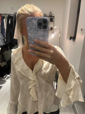 Zara blus med volang  - Säljer en krämvit blus med v-ringning och volangdetaljer längs halsringningen och framtill. Blusen har ett subtilt blommönster i tyget och vida, långa ärmar med volang vid ärmslut. Materialet är lätt och har en fin struktur som ger en lyxig känsla.