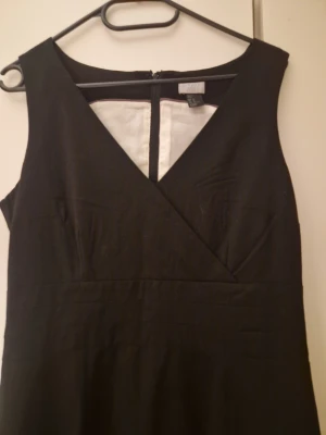 Svart omlottklänning Modern Classic - Svart ärmlös klänning från Modern Classic i omlottmodell med v-ringning. Klänningen har en stilren och tidlös design, dragkedja bak och är fodrad med ljus insida. Perfekt för dig som gillar klassiska och enkla plagg.