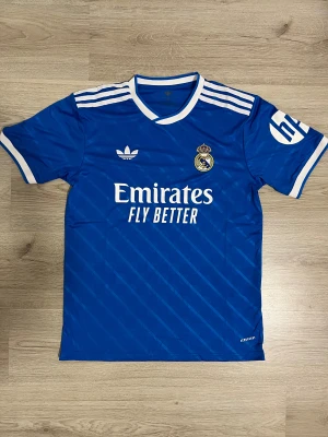 Blå Real Madrid t-shirt Adidas M - Säljer en blå Real Madrid t-shirt från Adidas med vita detaljer och ränder på axlarna. Tryck med 'Emirates Fly Better' på bröstet och klubbmärke. T-shirten har korta ärmar, v-ringad krage och diskret randigt mönster i tyget. Perfekt för fotbollsfans!