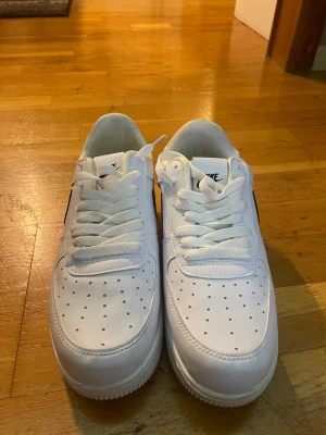 Nike Air Force 1 vit/svart sneakers - Nike Air Force 1 sneakers i klassisk vit med svarta detaljer och swoosh. Lågt skaft, perforerad tå och platt sula. Skorna har snörning och är tillverkade i skinn med syntetfoder. Perfekt för dig som gillar stilrena och tidlösa sneakers med streetkänsla.