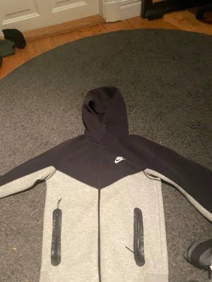 Nike tvåfärgad hoodie med dragkedja - Nike hoodie i svart och ljusgrått med hel dragkedja och två fickor med svarta detaljer. Klassisk Nike-logga på bröstet och stor huva. Materialet är mjukt och bekvämt, perfekt för chill eller träning. Snygg kontrast mellan färgerna och stilren design. Lite sol bränd på luvan men inget man tänker på.
