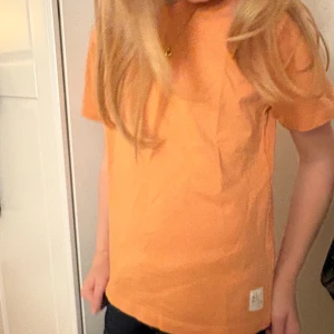 Orange t-shirt - Snygg orange t-shirt från Minimarket i mjuk bomull. Klassisk rund hals och korta ärmar, med en liten etikett nertill. Perfekt för dig som gillar färg och enkel stil. Passar dig som vill ha en chill och bekväm look. Otroligt liten storlek men passar på modellen som är 140cm