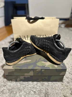 Valentino Rockrunner svarta sneakers - Svarta Valentino Rockrunner sneakers med coola nitar på sidorna och häl. Snygg mix av skinn och mocka, beige sula och klassisk snörning. Perfekt för dig som gillar edgy designerstil och vill sticka ut med detaljer. Kommer med originalkartong med camouflage-mönster. Box medföljer ej men kan förhandlas riktigt bra skick