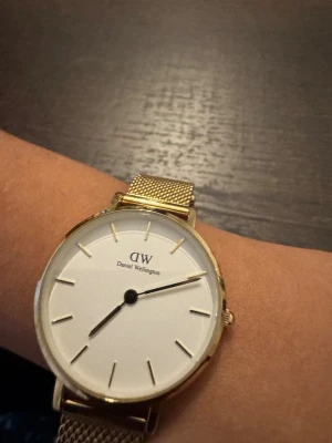 Guldfärgad klocka från Daniel Wellington - Snygg och stilren klocka från Daniel Wellington med vit urtavla och guldfärgat mesharmband i metall. Minimalistisk design med tunna guldmarkeringar och DW-logga på urtavlan. Perfekt accessoar för dig som gillar klassisk och clean look.