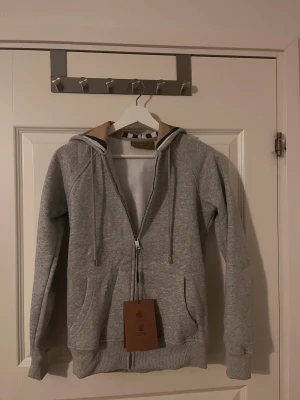 Burberry hoodie - Snygg grå hoodie från Burberry med klassiskt rutigt foder i huvan. Tröjan har dragkedja framtill, snörning vid huvan och två fickor. Perfekt för dig som gillar stilrena och exklusiva streetwear-plagg med en twist av lyx. Aldrig använd