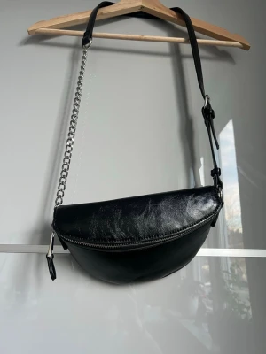 Svart axelväska från Gina Tricot - Snygg svart axelväska från Gina Tricot i glansigt syntetmaterial med silverfärgad kedja och justerbart axelband. Väskan har en rundad form och dragkedja upptill. Perfekt för att bära det viktigaste och ger en cool vibe till din outfit.