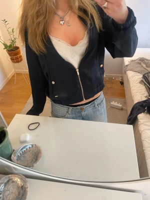 Svart bomberjacka Holly & Whyte M - Svart bomberjacka från Holly & Whyte by Lindex i storlek M. Jackan har dragkedja framtill, två fickor med guldfärgade knappar och elastisk midja. Materialet känns lätt och skönt, perfekt för att slänga på sig över en t-shirt eller linne.