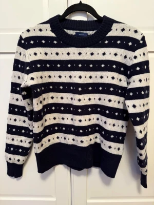 Randig stickad tröja från GANT - Säljer en snygg stickad tröja från GANT med marinblå och vita ränder samt diskret mönster. Tröjan har rund hals, ribbade muddar och långa ärmar. Perfekt för kyliga dagar och passar dig som gillar klassisk stil.