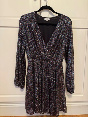 Glittrig kortklänning från Kilky Paris - Säljer en glittrig svart kortklänning från Kilky Paris i storlek M/L. Klänningen har långärmade ärmar, djup v-ringning och är täckt av färgglada paljetter som ger ett riktigt party-vibe. 