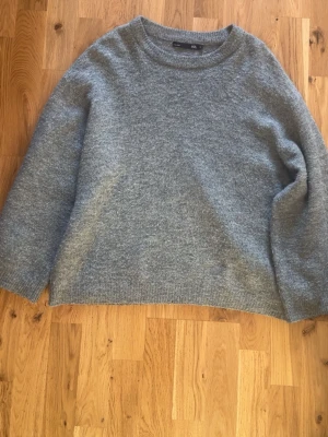 Grå stickad tröja  - Mysig grå stickad tröja med rund hals och långärmad design. Perfekt för kyliga dagar och enkel att matcha med jeans eller kjol. Tröjan har en loose passform och ribbade muddar vid ärmslut och nederkant.