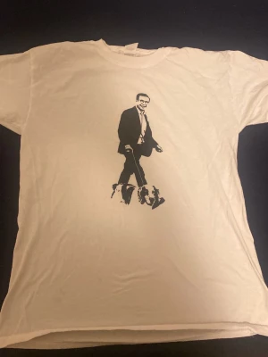 Vit t-shirt med svart tryck av man och hund - Vit t-shirt i bomull med ett stort svart tryck på framsidan som föreställer Ulf Kristersson i kostym som gå med en hund. Klassisk passform och rund halsringning. Perfekt för dig som gillar unika grafiska motiv.