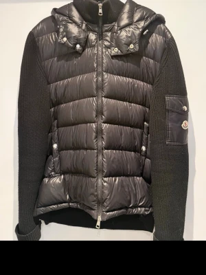 Svart Moncler pufferjacka med huva - Säljer en svart pufferjacka från Moncler med stickade ärmar och vadderad kropp. Jackan har dragkedja framtill, fickor med tryckknappar och en avtagbar huva. Moncler-logga på ärmen och snygga detaljer som ribbade muddar. Perfekt för kyliga dagar.
