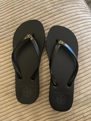  Tory Burch - Snygga svarta flipflops från Tory Burch med guldigt logodetalj på remmen. Skorna har en klassisk form med rund tå och platt sula, tillverkade i syntetmaterial. Perfekta för sommardagar och strandhäng. Storlek 36/37